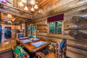A log cabin interior with a wooden dining table, chairs, a kitchen area, and a large window. - Cozy Log Cabin Retreat, Sleeps 10・Hot Tub・Firepit (Sevierville)