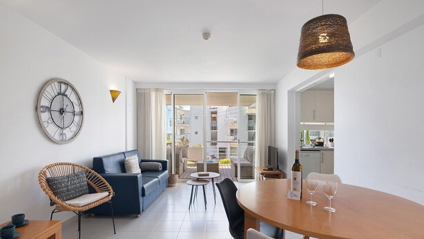 A modern living room with a sofa, a dining table, a wicker chair, a clock, and a view through a glass door to an outdoor balcony.