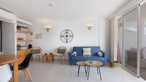 A modern living room with a blue sofa, two wicker chairs, a wooden dining table, and a round coffee table.