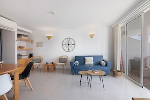 A modern living room with a blue sofa, two wicker chairs, a wooden dining table, and a round coffee table.