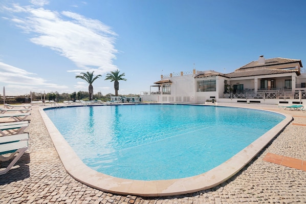 A swimming pool with lounge chairs, a building with a balcony, and palm trees in the background.