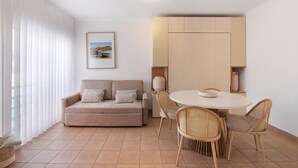 Ein modernes Wohnzimmer mit einer Couch, einem runden Esstisch, Korbstühlen und einem großen Fenster mit Jalousien.