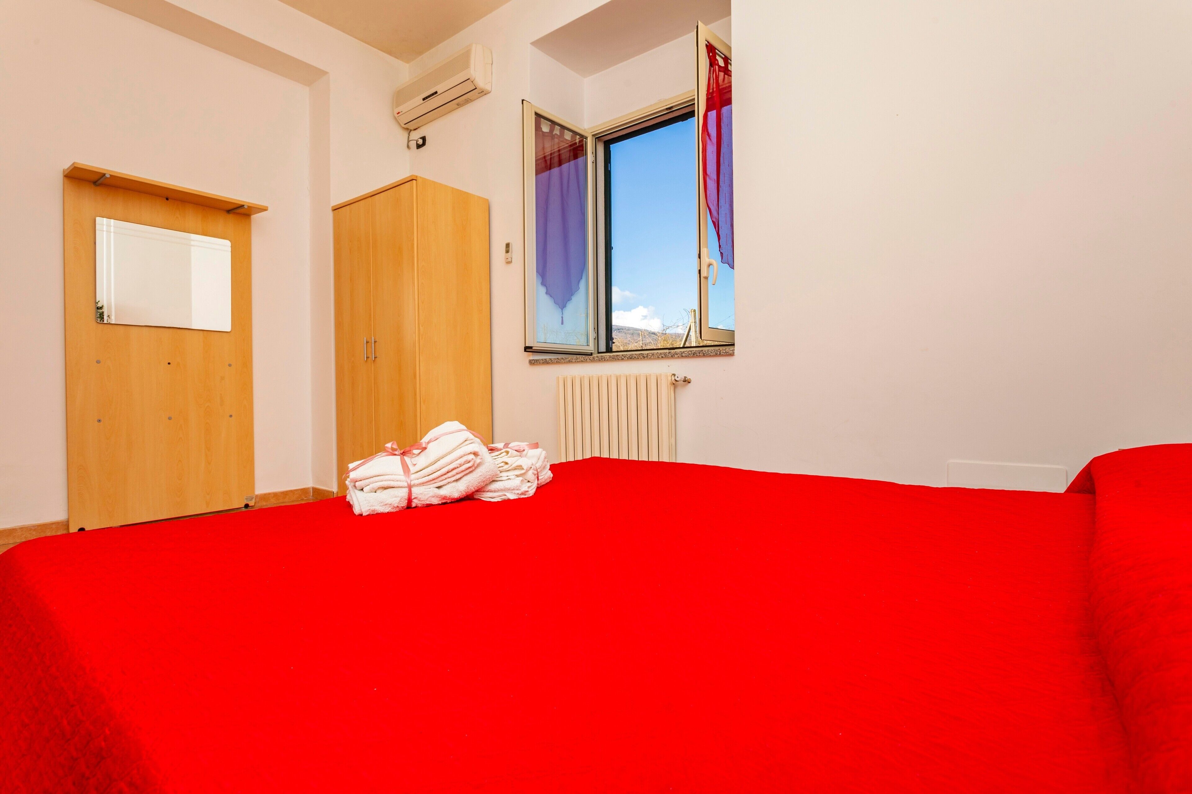 Une chambre avec un lit rouge, une armoire en bois et une fenêtre avec des rideaux.