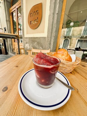 Una mesa con un vaso de bebida roja, un plato blanco con borde azul y una cesta de pan.