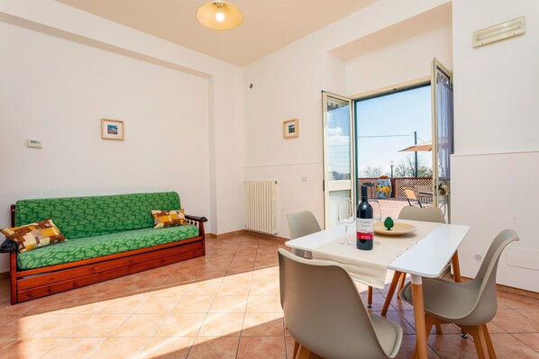 A room with a green sofa, a dining table with chairs, a bottle of wine, and a view through a glass door.