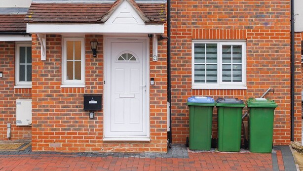 A two-story brick house with a white front door, two windows on the ground floor, and a small porch. There are green bins outside the house.