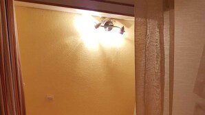 Una habitación pequeña con una cama individual, una colcha blanca sencilla, una mesita de noche y una lámpara fijada a la pared.