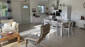 Amplia sala de estar con sofá, una mesa de centro y una mesa de comedor con sillas. Hay una cocina con refrigerador, estufa y lavaplatos.