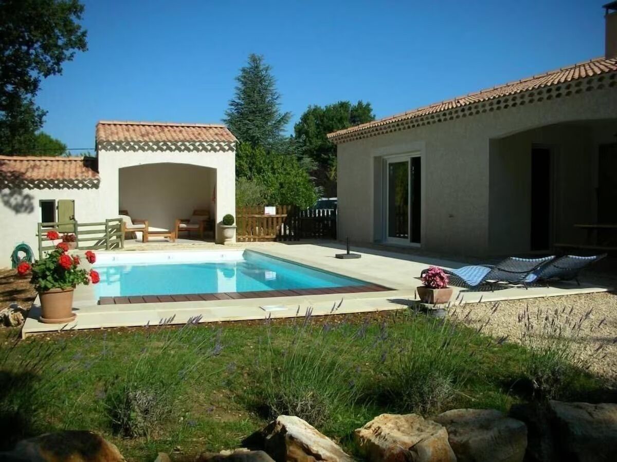 A swimming pool with a tiled deck, surrounded by a garden with potted plants and a wooden fence.