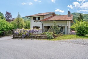 A two-story house with a red-tiled roof, a fenced garden with purple flowers, and a mountainous backdrop.