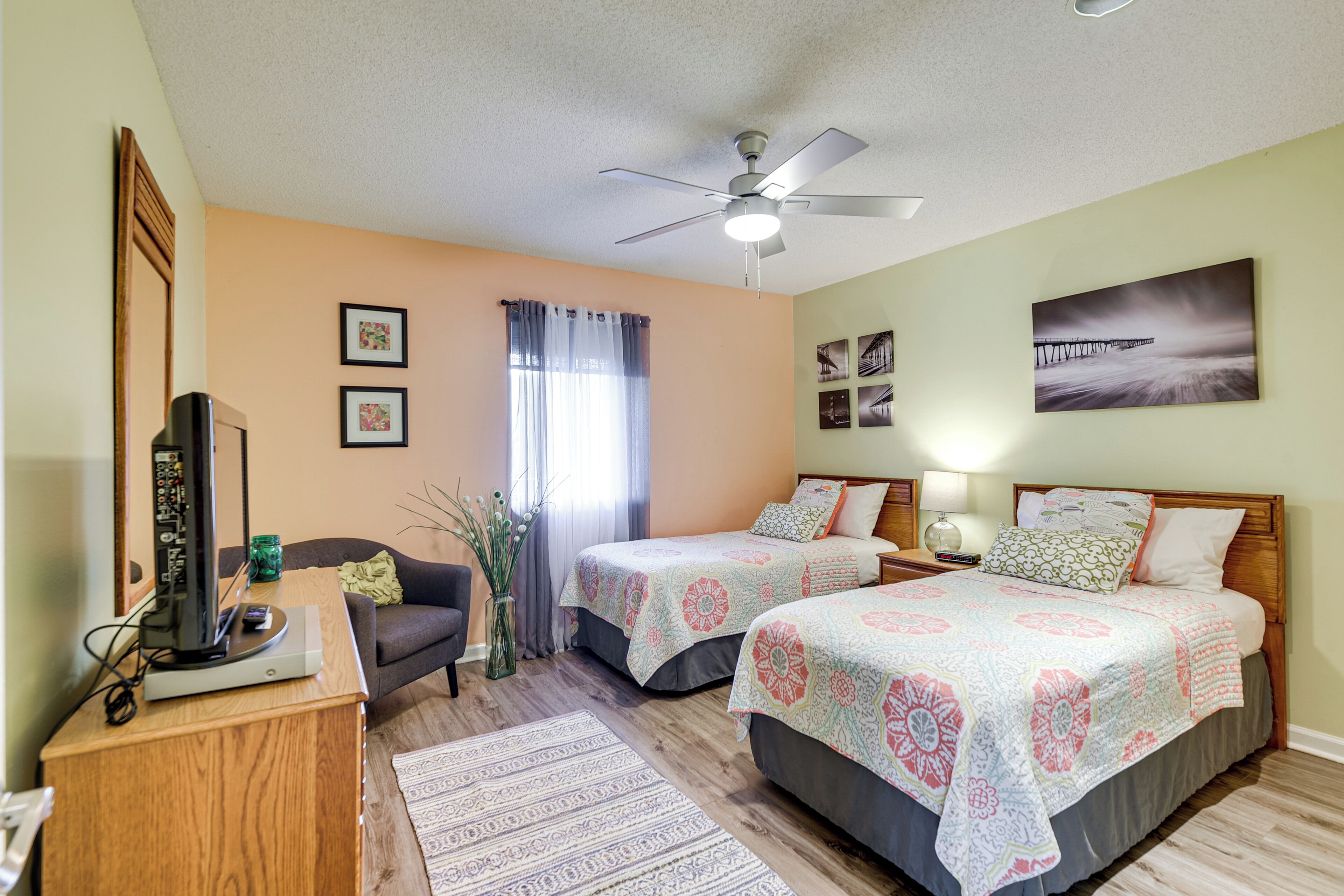 A bedroom with two beds, a ceiling fan, a desk with a computer, and a window with curtains.