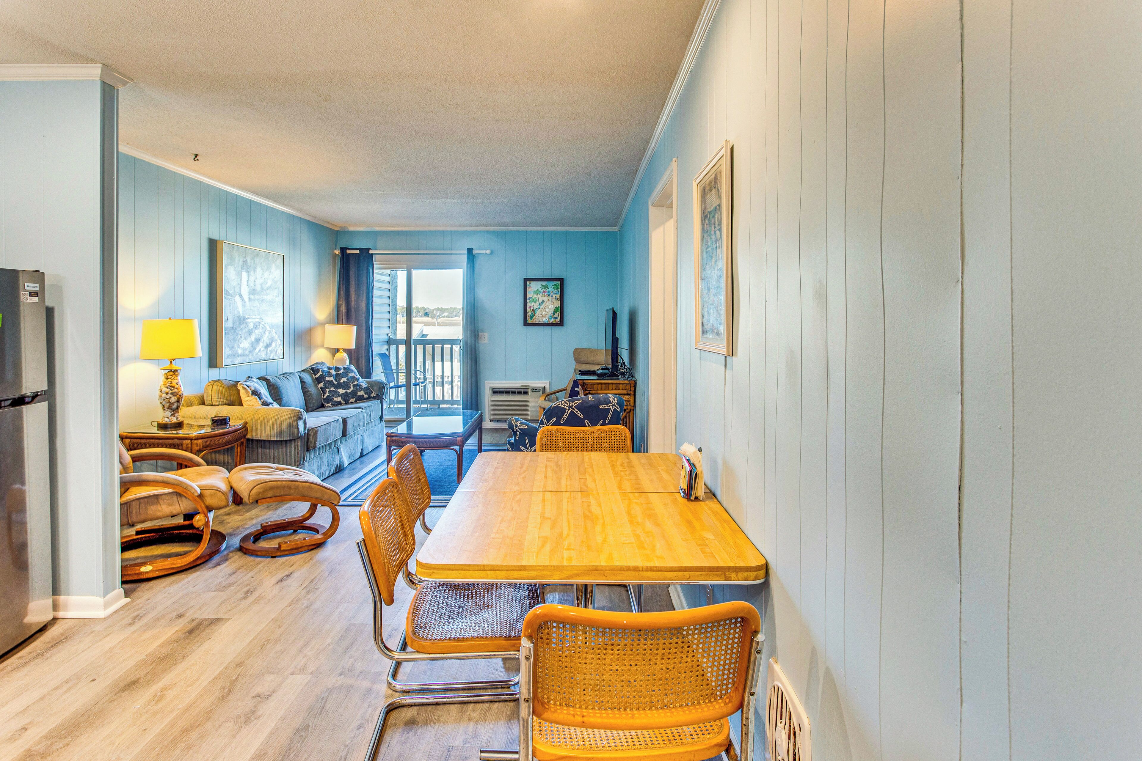 A dining area with a wooden table and wicker chairs, a sofa, and a small kitchen area with a refrigerator.