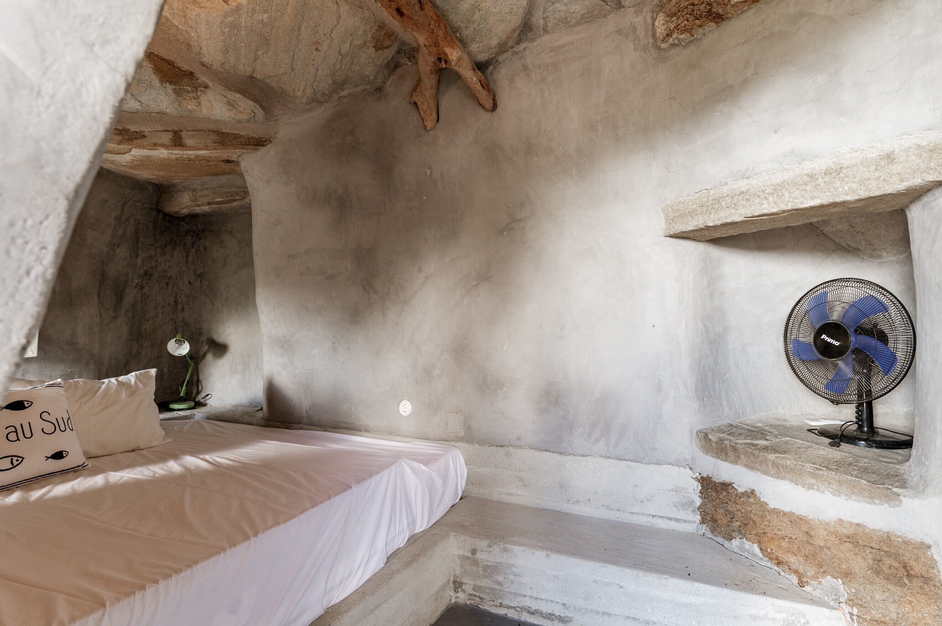 Una habitación con cama, mesita de noche, lámpara y ventilador de pared.