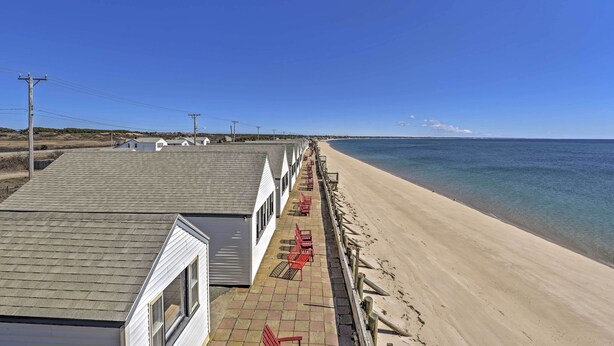 A beachfront property with a row of white cabins, a sandy beach, and a clear blue sky. - Ocean-view Cape Cod Condo in Truro! (North Truro)