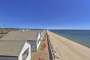 A beachfront property with a row of white cabins, a sandy beach, and a clear blue sky. - Ocean-view Cape Cod Condo in Truro! (North Truro)