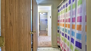 A hallway with a wooden door, a colorful curtain, and a carpeted area leading to a room with a bed and a ceiling light. - Ocean-view Cape Cod Condo in Truro! (North Truro)