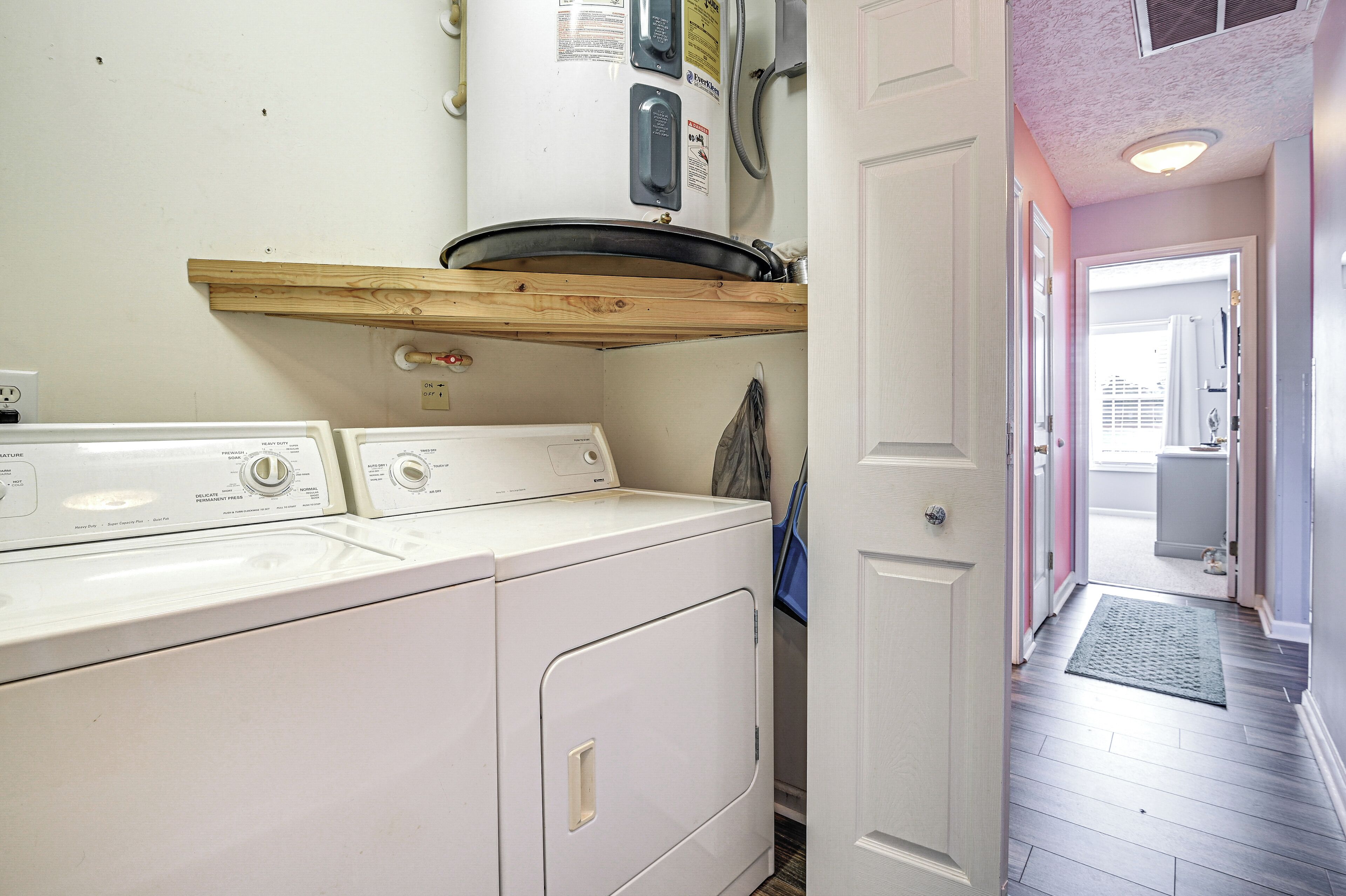 A laundry area with two front-loading washing machines, a wooden shelf above, and a door leading to a hallway with a rug.