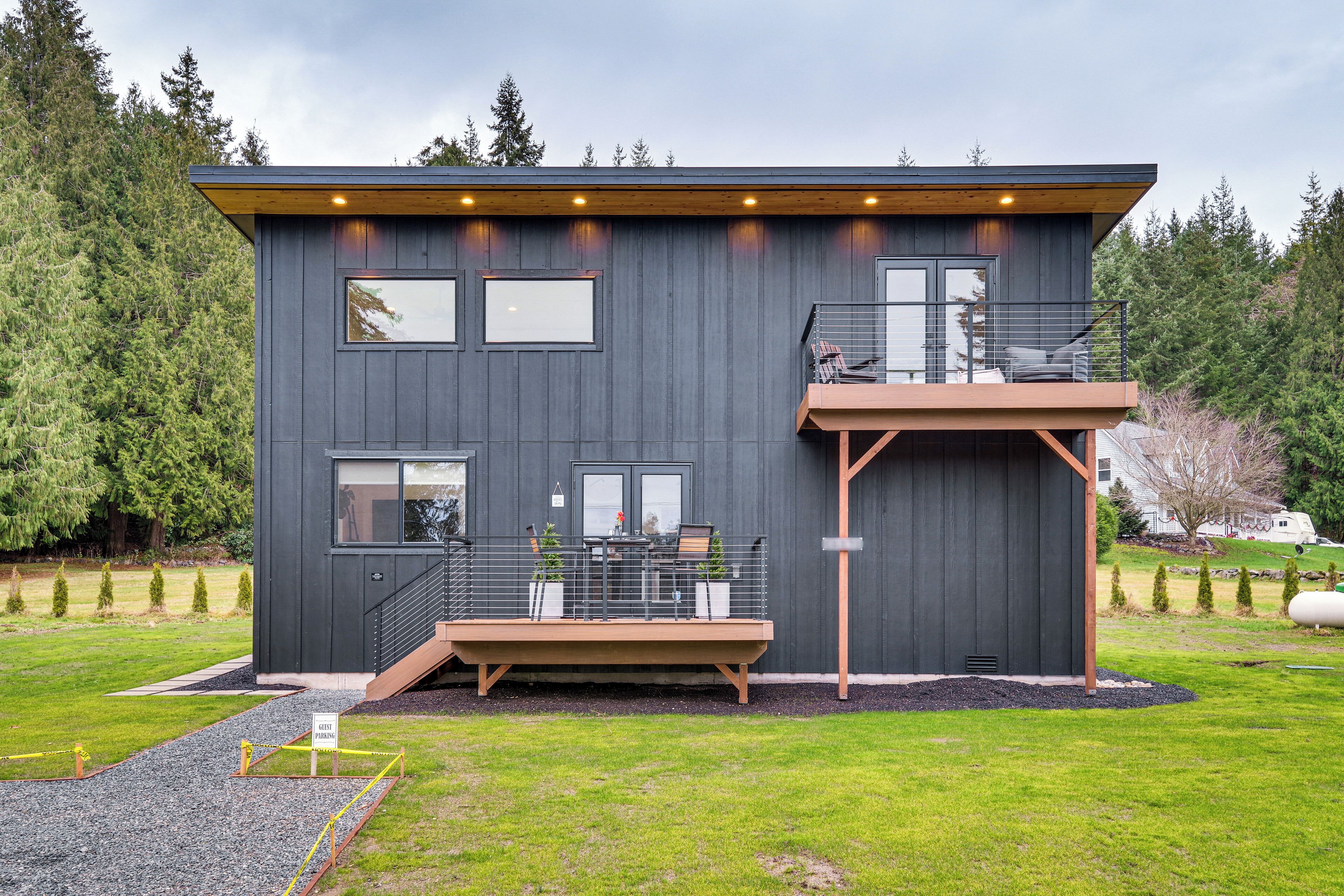 A modern two-story house with a balcony, a deck, and a view of the surrounding forest.