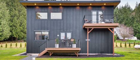 A modern two-story house with a balcony, a deck, and a view of the surrounding forest.