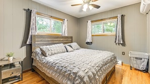 A bedroom with a bed, a ceiling fan, two windows with curtains, a nightstand with a plant, and a laundry cart. - Canal-front Cabin: Dock & Fire Pit in New Lisbon (New Lisbon)
