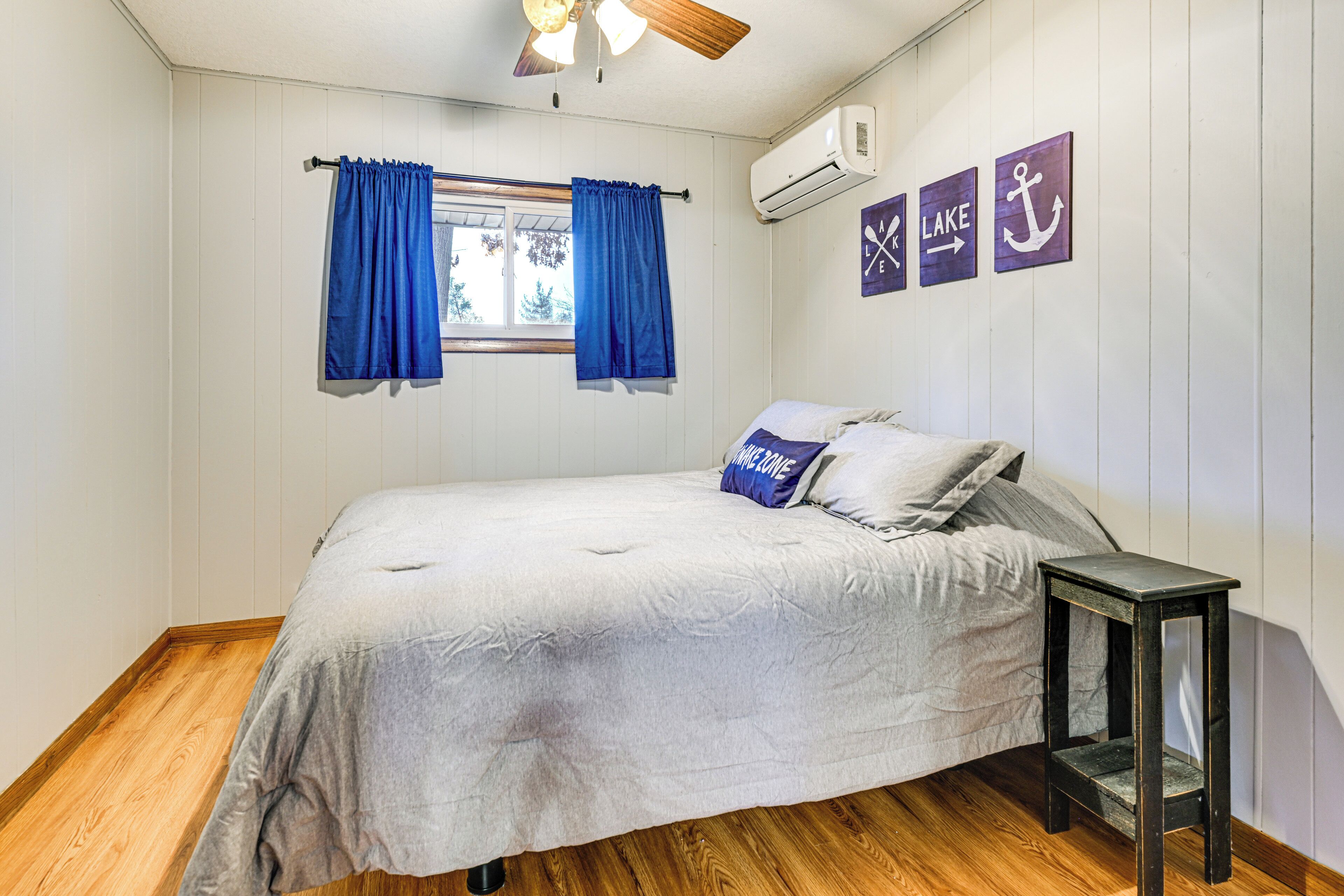 Une chambre à coucher avec un lit, une fenêtre ornée de rideaux bleus, une petite table et des éléments de décoration murale, notamment un panneau représentant une ancre et le mot « LAKE ».