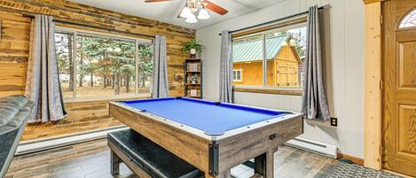 A pool table in a room with wooden walls, a ceiling fan, and large windows.