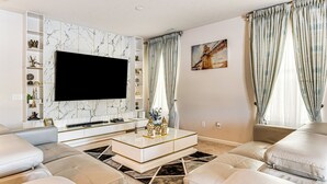 Ein modernes Wohnzimmer mit Flachbildfernseher, Wand mit Marmoroptik, Glastisch und großem Fenster mit Vorhängen.