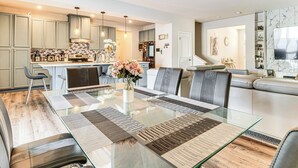 Eine moderne Küche mit einer zentralen Kochinsel, ein Essbereich mit einem Glastisch und ein Wohnbereich mit Sofa und Fernseher.