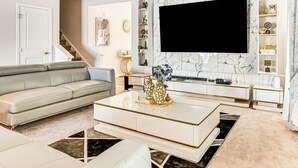 Ein modernes Wohnzimmer mit einem großen Flachbildfernseher, einem grauen Sofa, einem weißen Couchtisch mit goldenen Akzenten und einer Wand mit Marmoroptik.