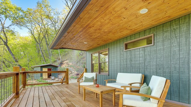 Une terrasse en bois avec des meubles d’extérieur, une vue sur la forêt et un bâtiment aux bardages verts.