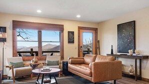 Un salon avec un canapé en cuir, deux fauteuils, une table basse et une vue sur les montagnes grâce à de grandes fenêtres.