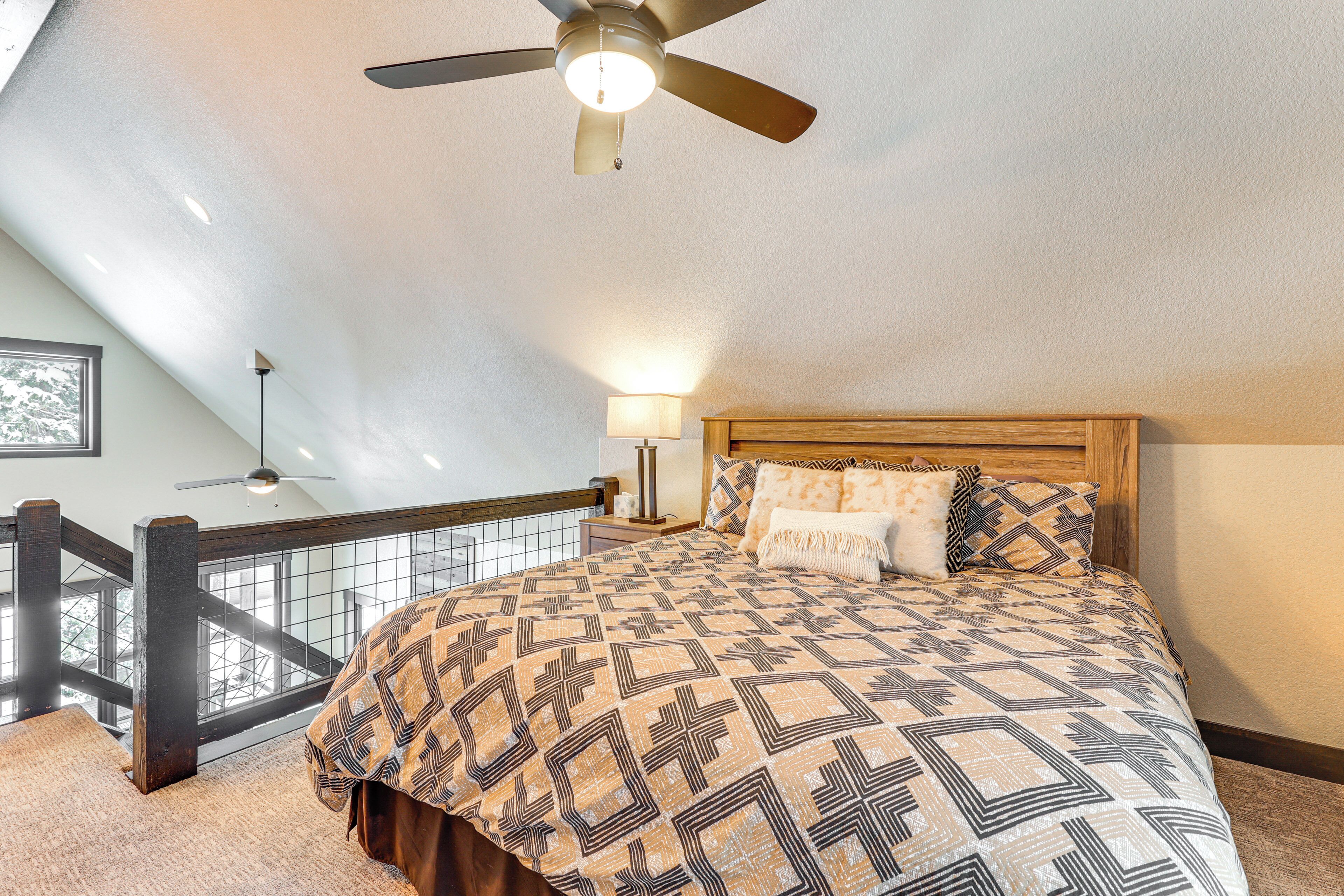A bedroom with a large bed, a ceiling fan, a nightstand, a lamp, and a view of a balcony.