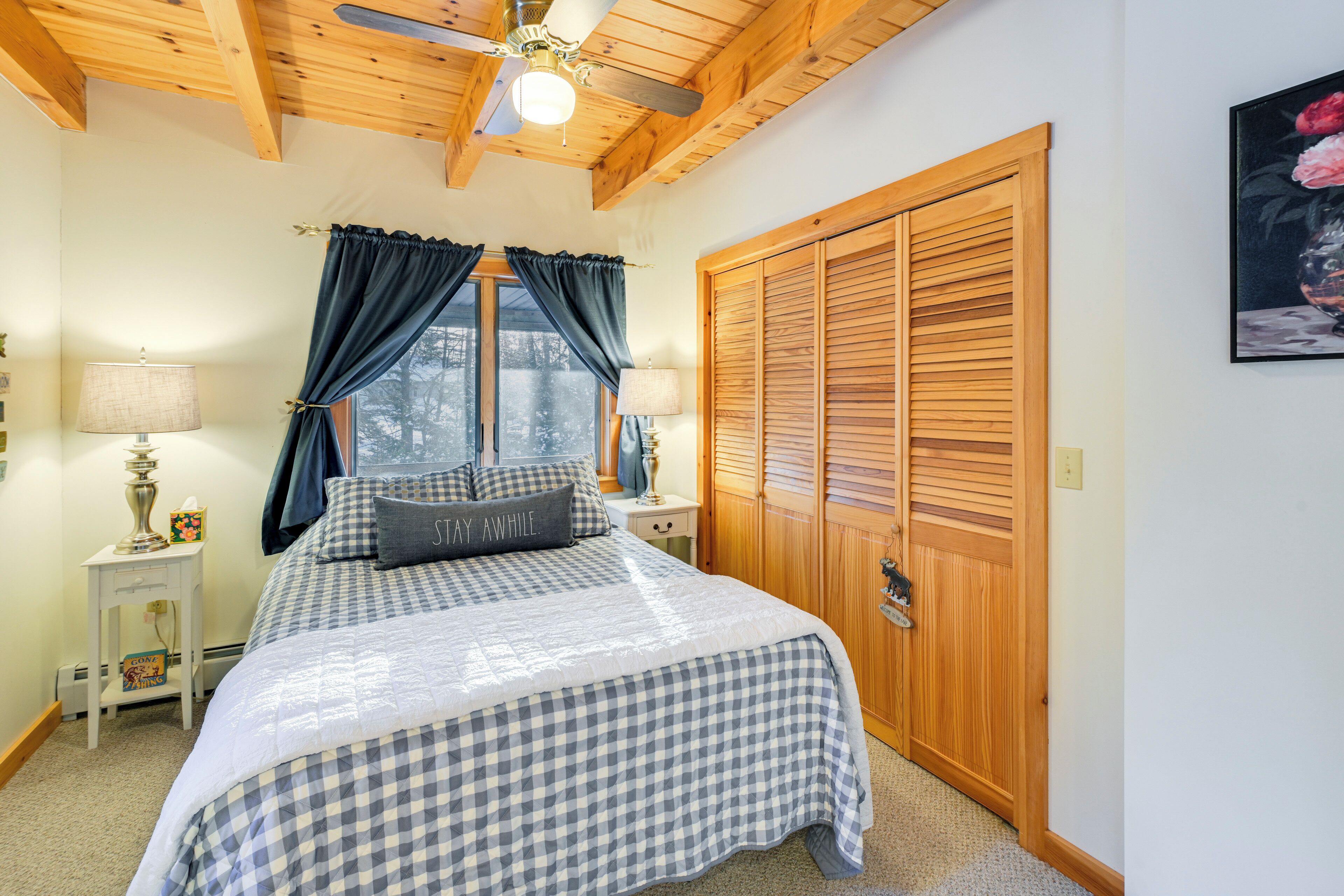 A bedroom with a checkered bedspread, a wooden wardrobe, a ceiling fan, and a window with curtains.