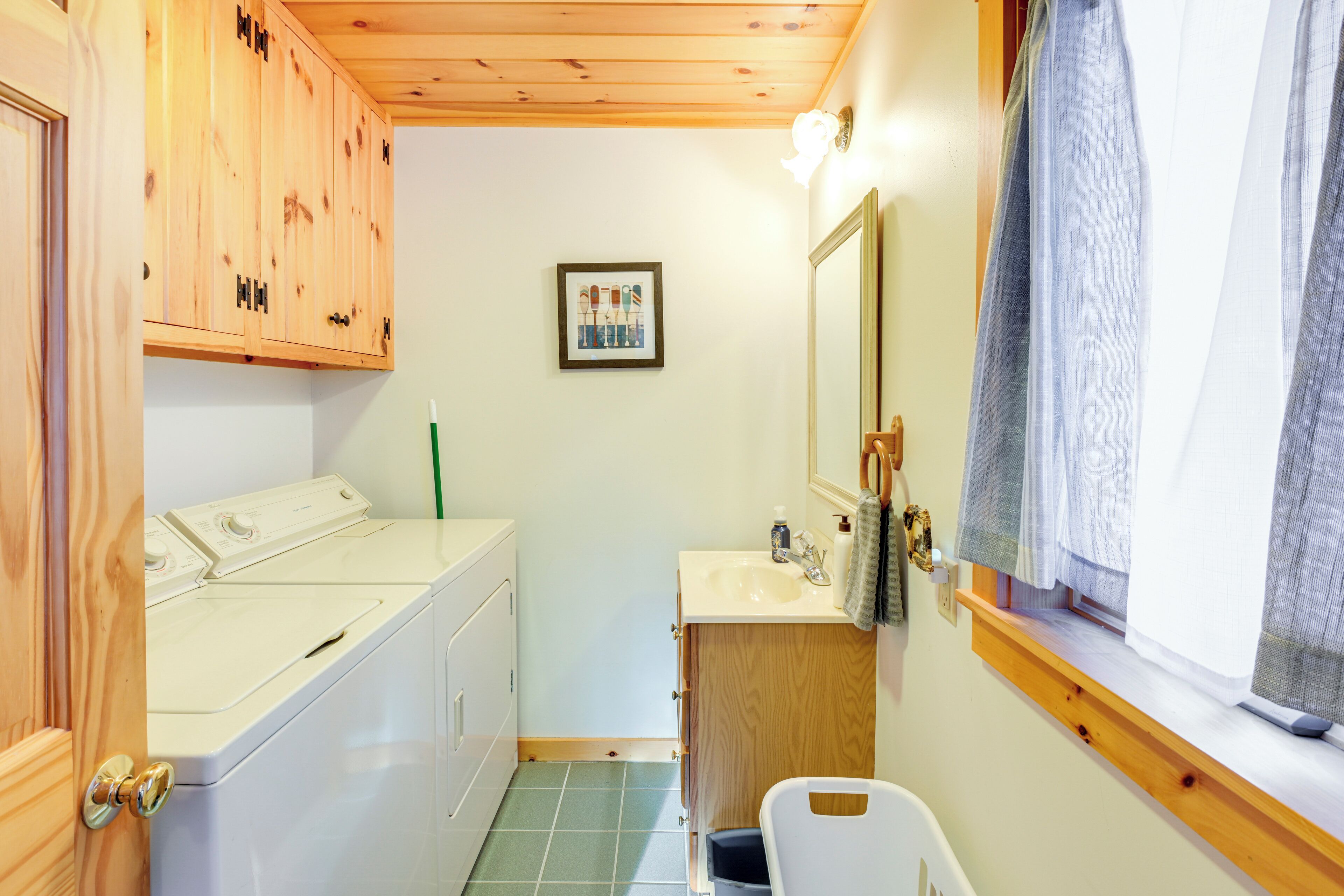 Un baño pequeño con armarios de madera, lavadora, lavamanos y ventana con cortinas.
