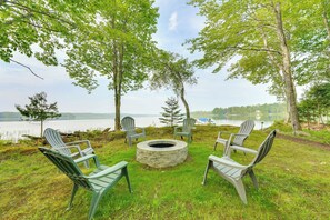 Un foyer circulaire en pierre, entouré de chaises Adirondack, installé sur une pelouse avec des arbres et un lac en arrière-plan.