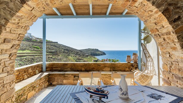 Arcada de pedra que dá acesso a um terraço com mesa, cadeiras e vista para o mar.