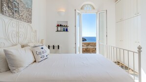 Quarto com cama, travesseiros, mesa de cabeceira, prateleira na parede com garrafas, um quadro e vista para o mar através de uma porta aberta.