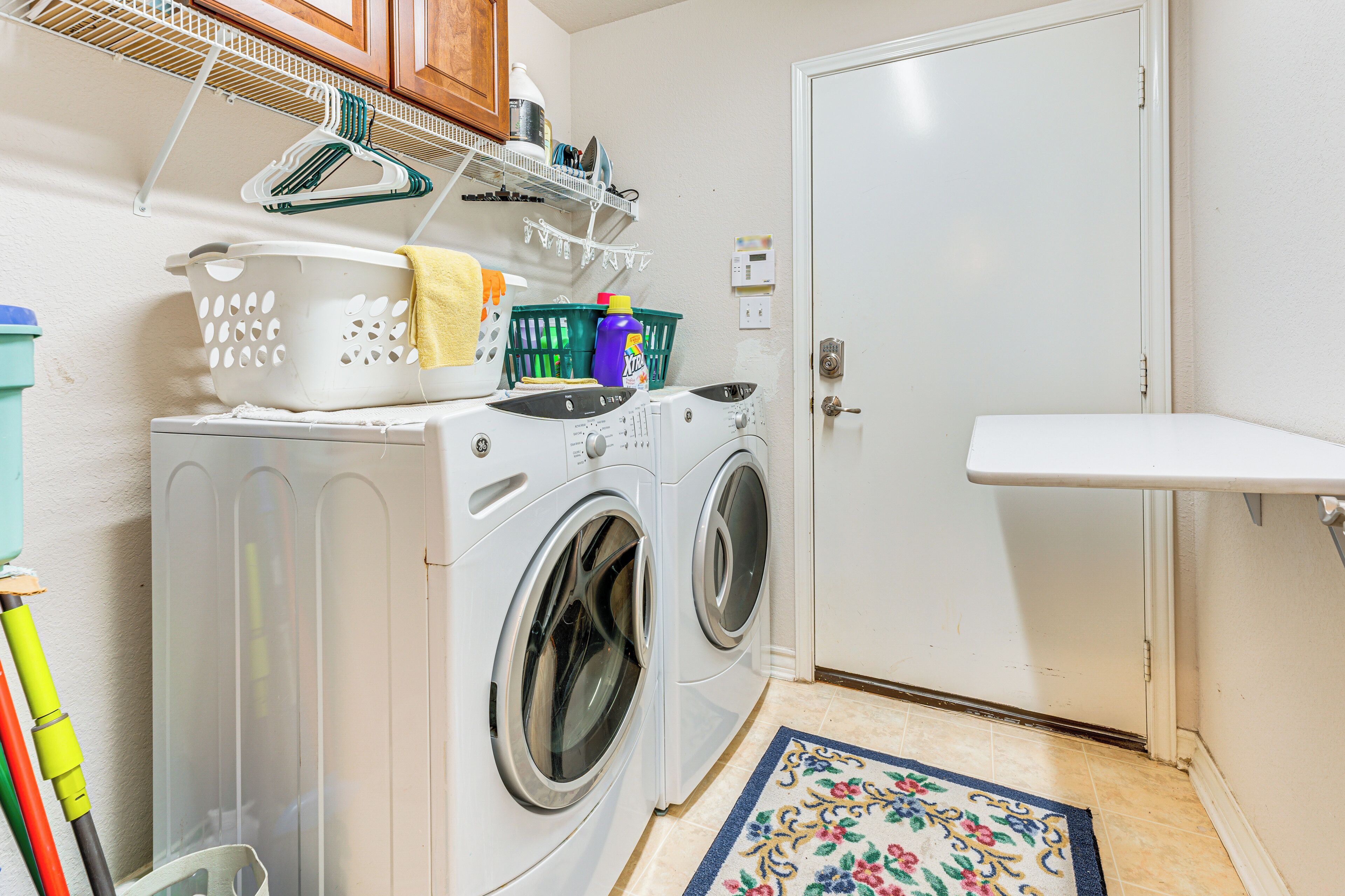 A laundry area with two front-loading washing machines, a drying rack, a basket, and a shelf with cleaning supplies.