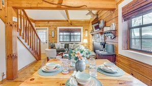 Interior de una cabaña de madera con una mesa de comedor lista para cuatro personas, una escalera y una sala de estar con sofá y chimenea.