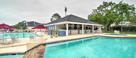 A swimming pool area with lounge chairs and umbrellas, a covered seating area, and a building with a sloped roof.