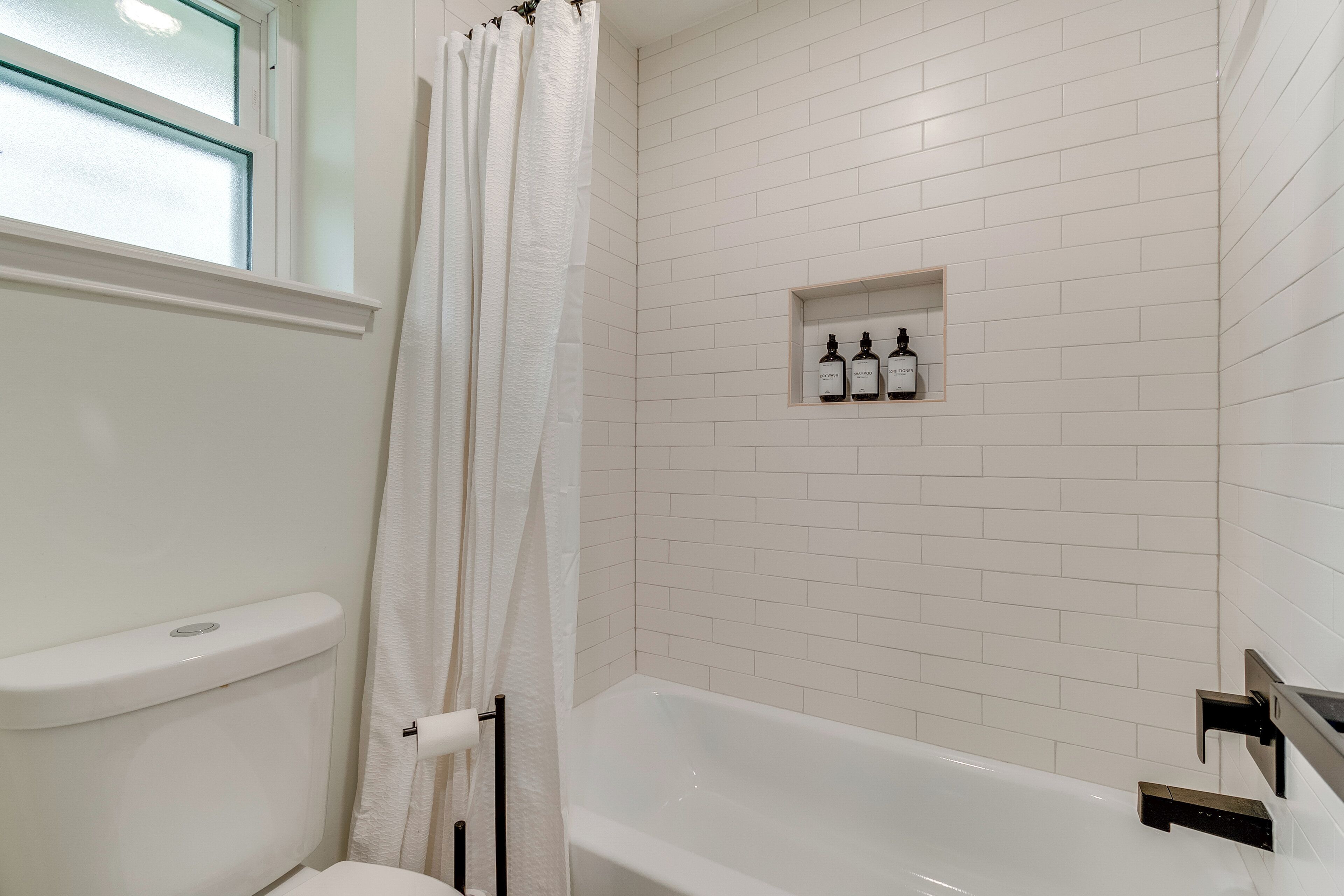 Une salle de bain avec une baignoire blanche, des toilettes et une fenêtre avec un vitrage dépoli. Il y a une étagère avec trois bouteilles accrochée au mur.