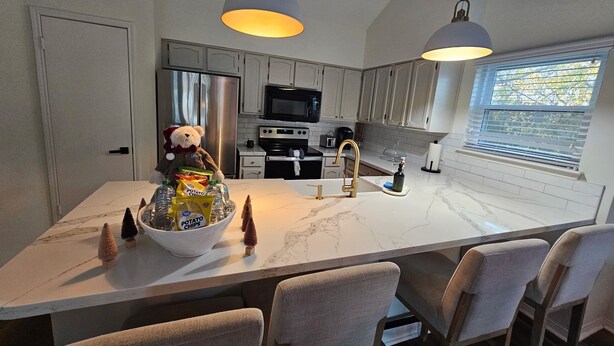 Une cuisine moderne avec des plans de travail en marbre blanc, un îlot central doté de tabourets, une décoration sur le thème d’un ours en peluche et une grande fenêtre avec des stores.
