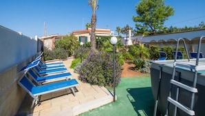 A rooftop pool area with blue lounge chairs, a palm tree, and a small garden with purple flowers.