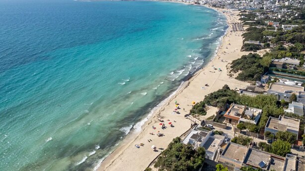 An aerial view of a coastal town with a sandy beach, clear blue waters, and residential buildings along the shore.