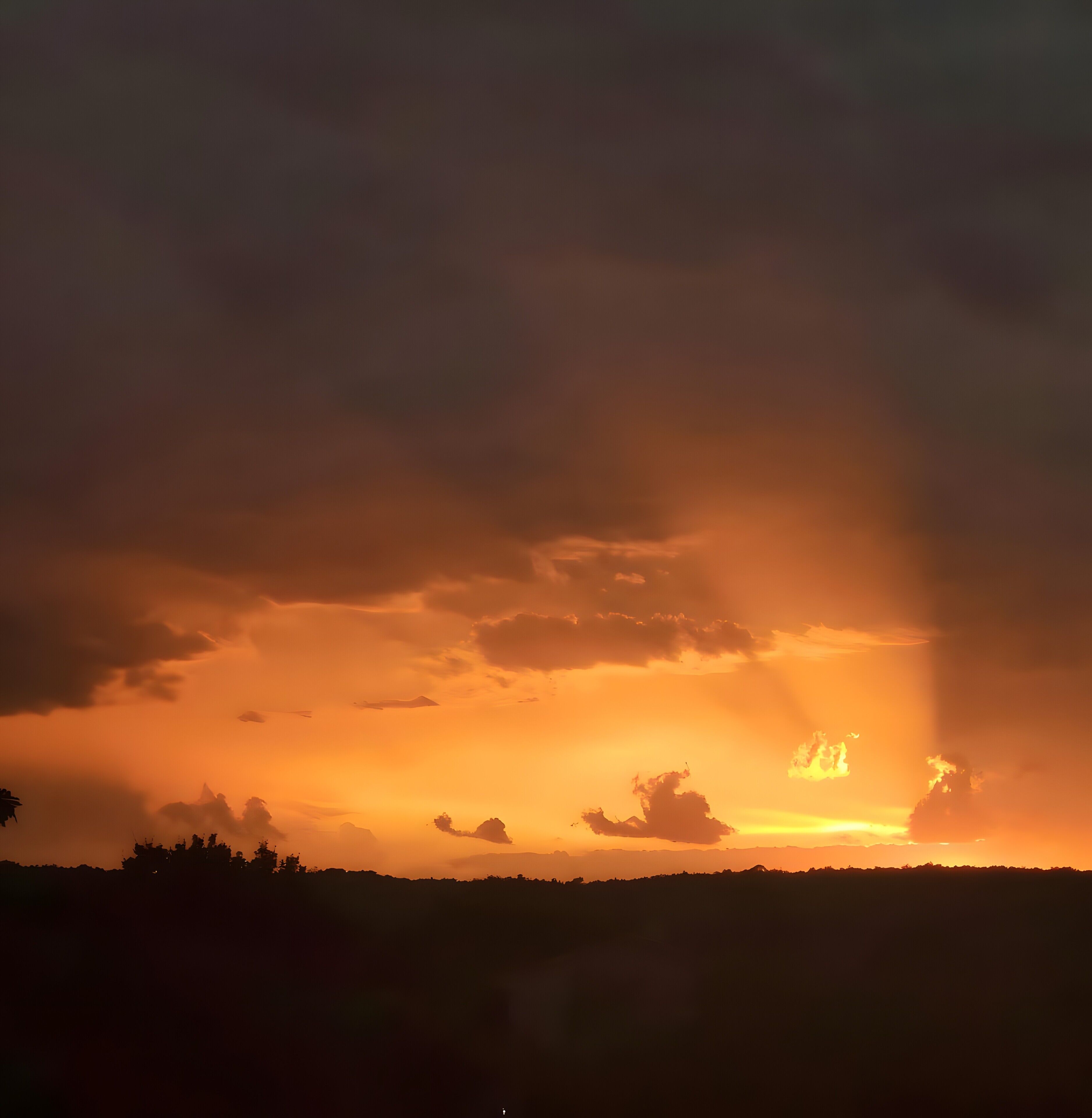 A sunset over a hilly landscape with silhouettes of trees and a distant mountain range.