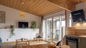 Ein modernes Wohnzimmer mit Holzboden, einem an der Wand befestigten Fernseher, einem Essbereich mit Tisch und Stühlen sowie einer Küche mit Herd.