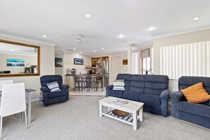 A living room with a sofa, armchair, and coffee table, a dining area with a white table and chairs, and a kitchen in the background. - Hidden Gem (Hamilton)