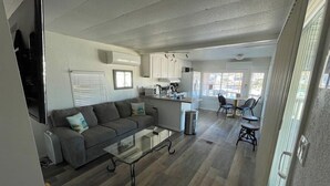 Un espacio habitable compacto con sofá, una mesita de centro, comedor con sillas, cocina con armarios y una ventana que proporciona luz natural.