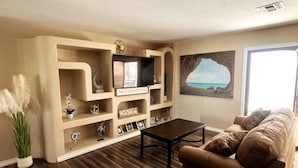 A living room with a brown leather sofa, a coffee table, and a wall-mounted TV. There are decorative items on shelves, and a painting of a beach scene is on the wall. - The Gentleman's Jack, Moovalya Keys Value (Parker)