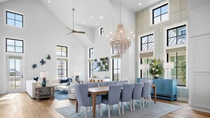 A spacious living area with a dining table, chairs, a chandelier, and a large window. - Beautifully Constructed Beach Home In Cinnamon Shore North. (Port Aransas)